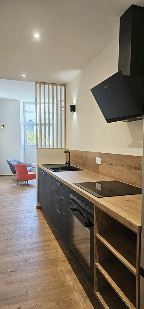 Cuisine moderne rénovée avec meubles noirs mats, plan de travail en bois clair, hotte design et séparation bois, projet d’architecte d’intérieur à Alès